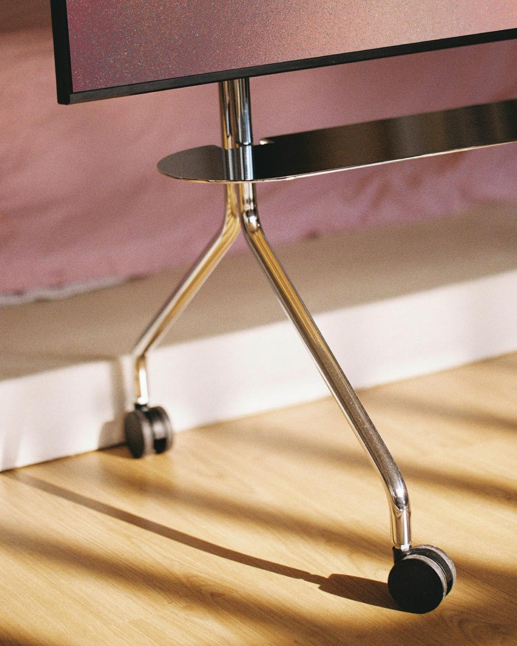 Close-up of the Moon Shelf in Chrome, showing its curved legs and black castor wheels on sunlit wood flooring