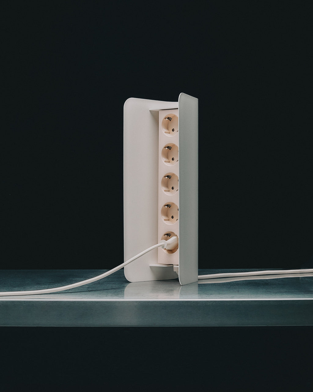 Power Shell in Pearl standing upright on a glass shelf, five-socket strip with plug inserted against dark studio backdrop