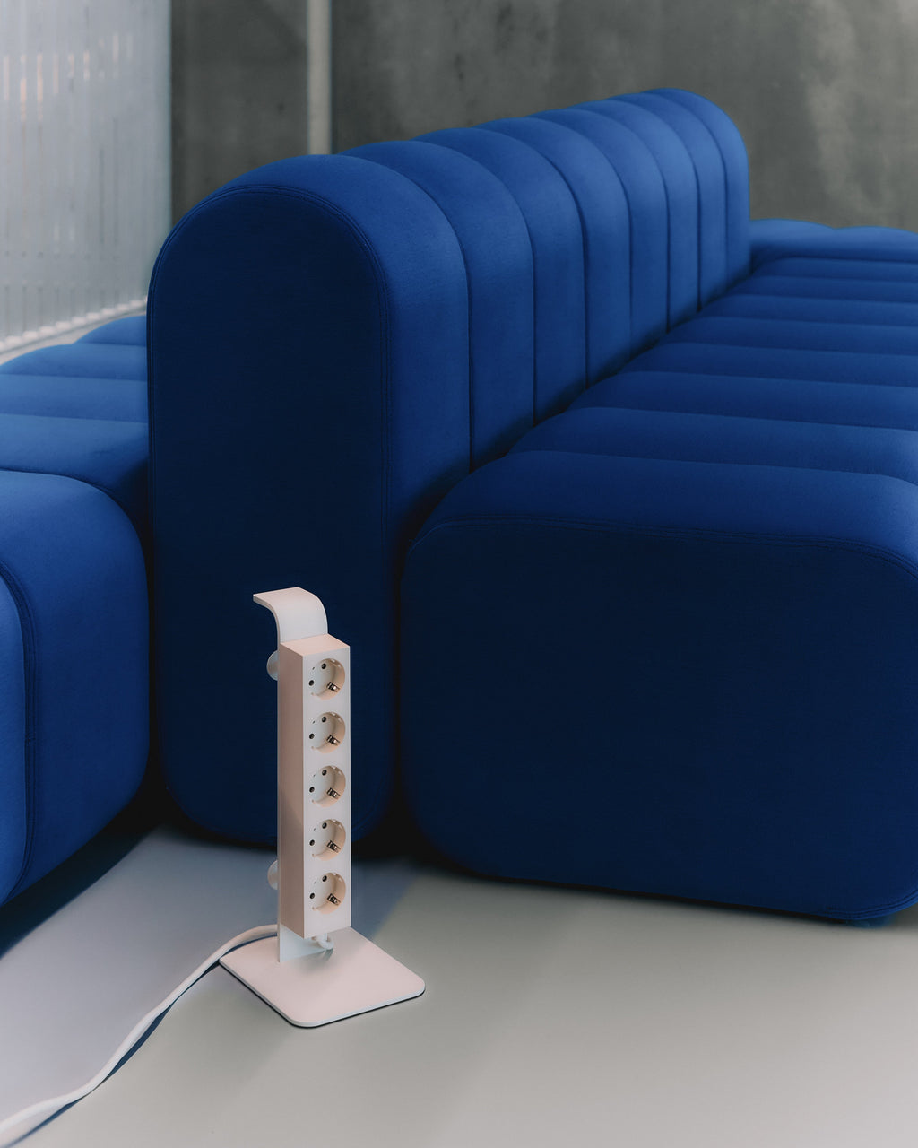 Pearl Power Tower with five vertical sockets standing on a grey floor beside a blue tufted modular sofa