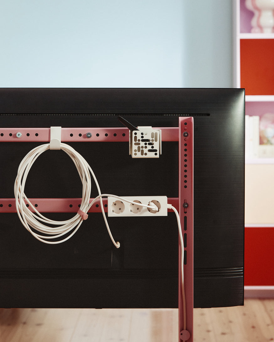Charcoal Remote Box Mount holding a remote behind a TV on a pink rail frame, with coiled cable and power strip