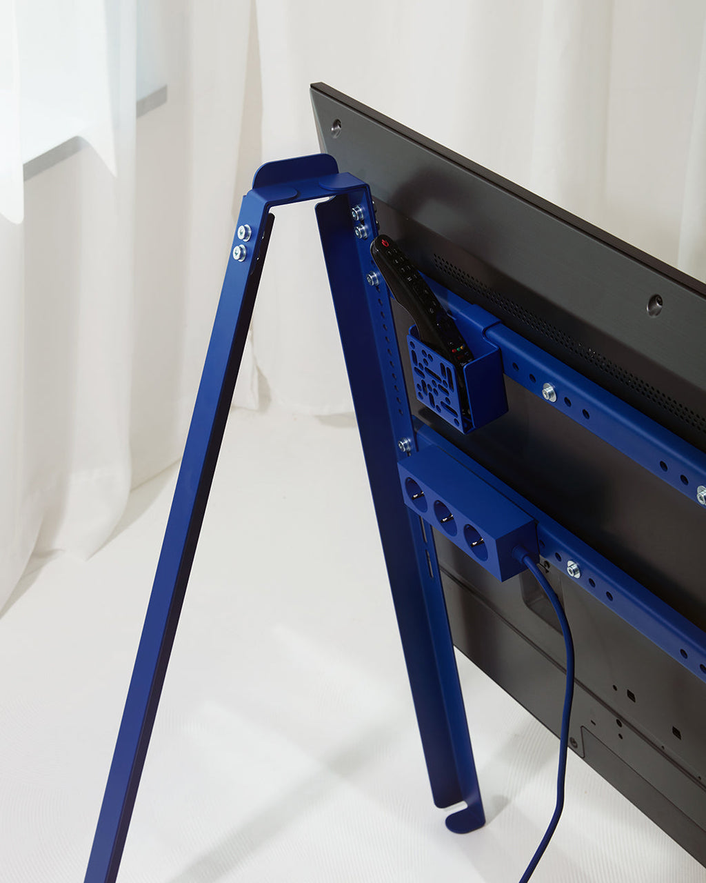 Ultra Marine Remote Box Mount holding a remote on a blue TV stand frame, rear view behind a mounted flatscreen