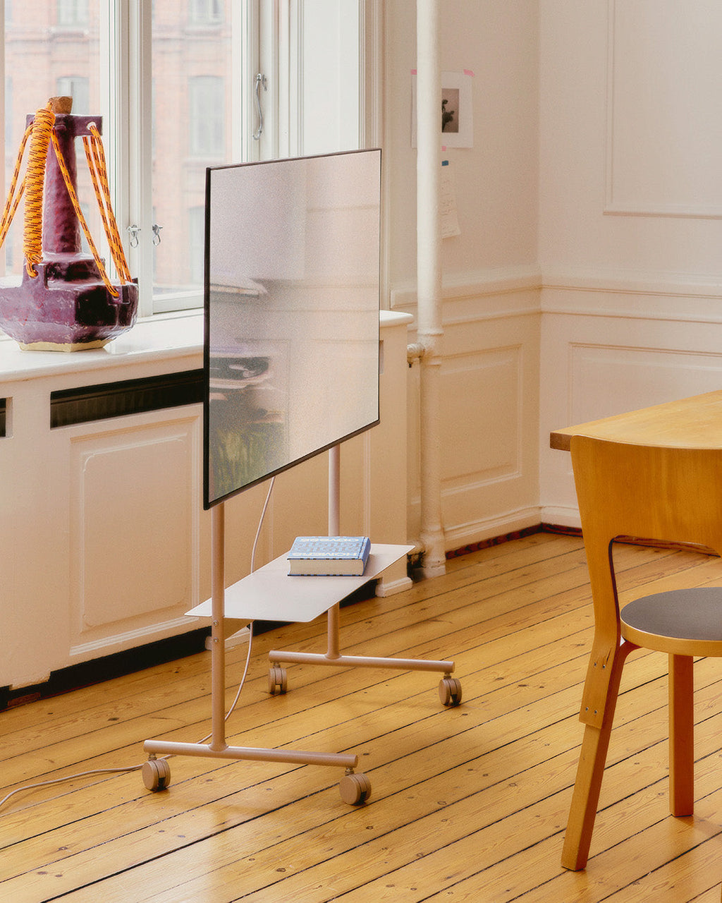 Rover stand in Charcoal on wheels holding a TV and book in a sunlit room with wood floors, angled side view