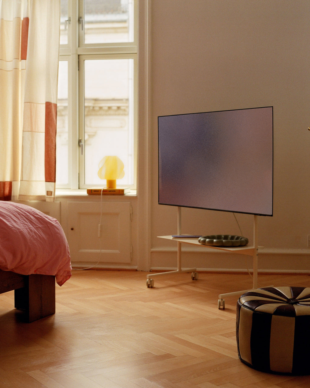 Rover TV stand in Pearl on wheels in a sunlit bedroom, holding a large flatscreen with books on its lower shelf