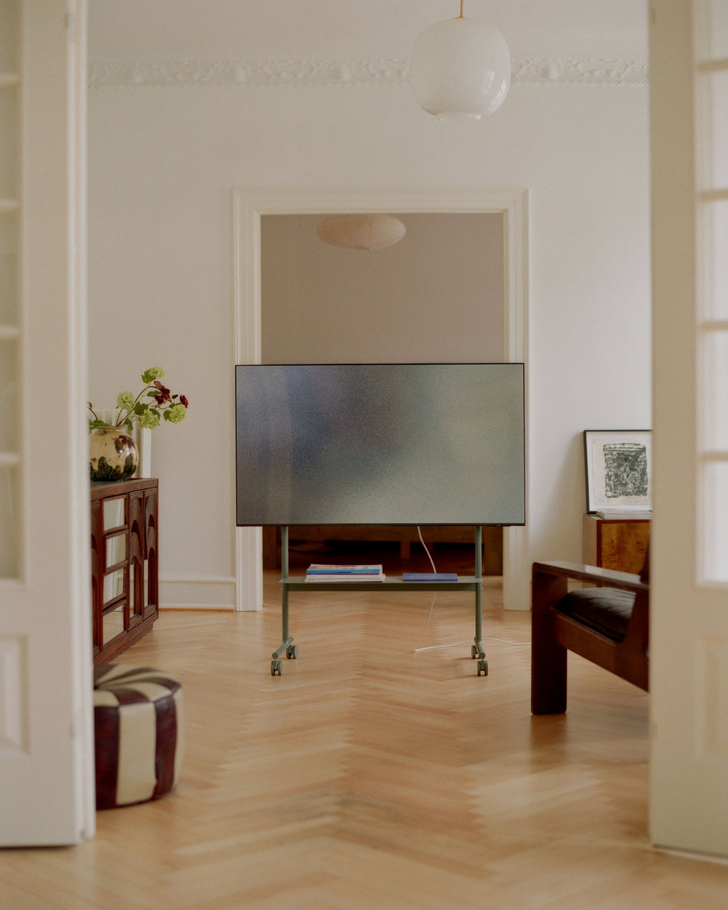 Rover mobile TV stand in Mossy Green holding a flat-screen in a herringbone-floor living room, magazines on lower shelf