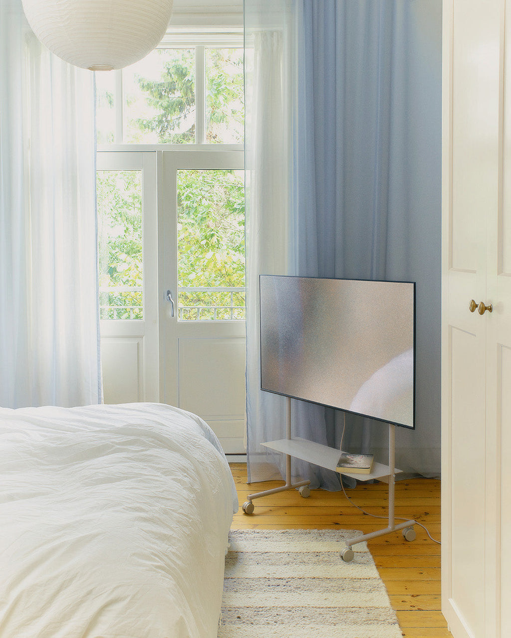 Rover TV stand in Oat on wheels holding a large TV in a bright bedroom, angled beside French doors and a striped rug