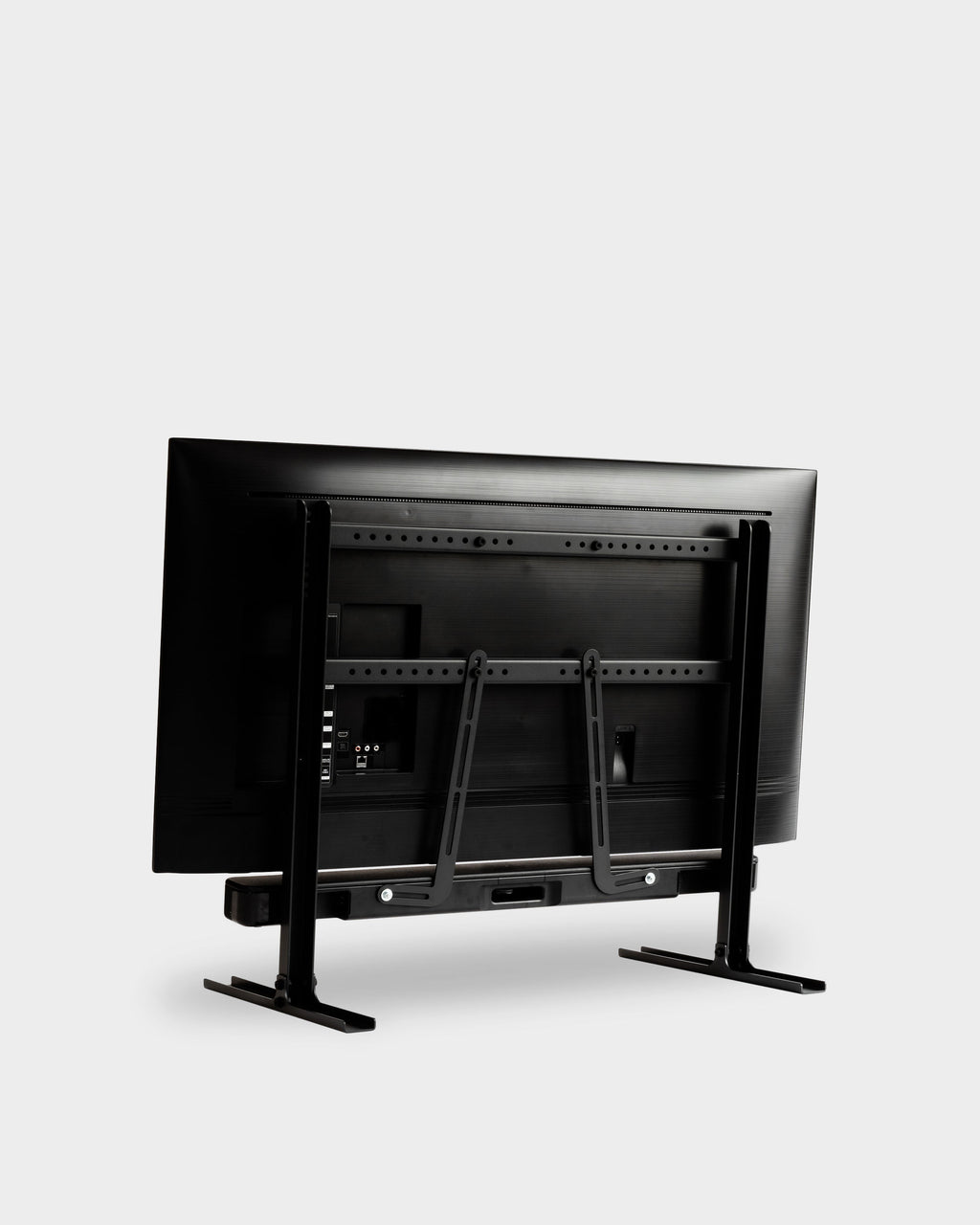 Charcoal Soundbar Bracket Set holding a soundbar behind a TV on tabletop stand, rear three-quarter studio view