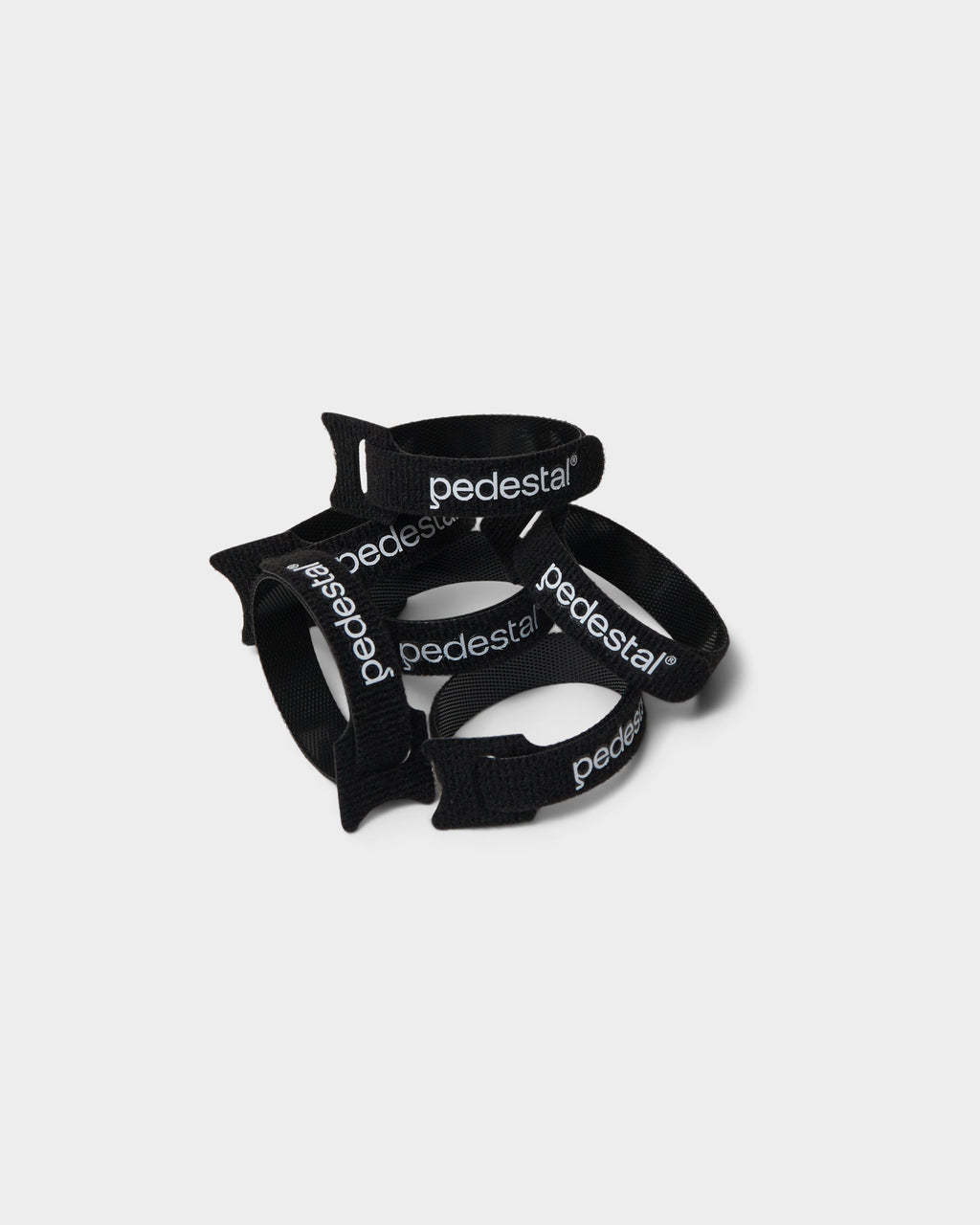 Stack of black Pedestal velcro cable ties with white logo print, loosely coiled in a studio packshot on light grey