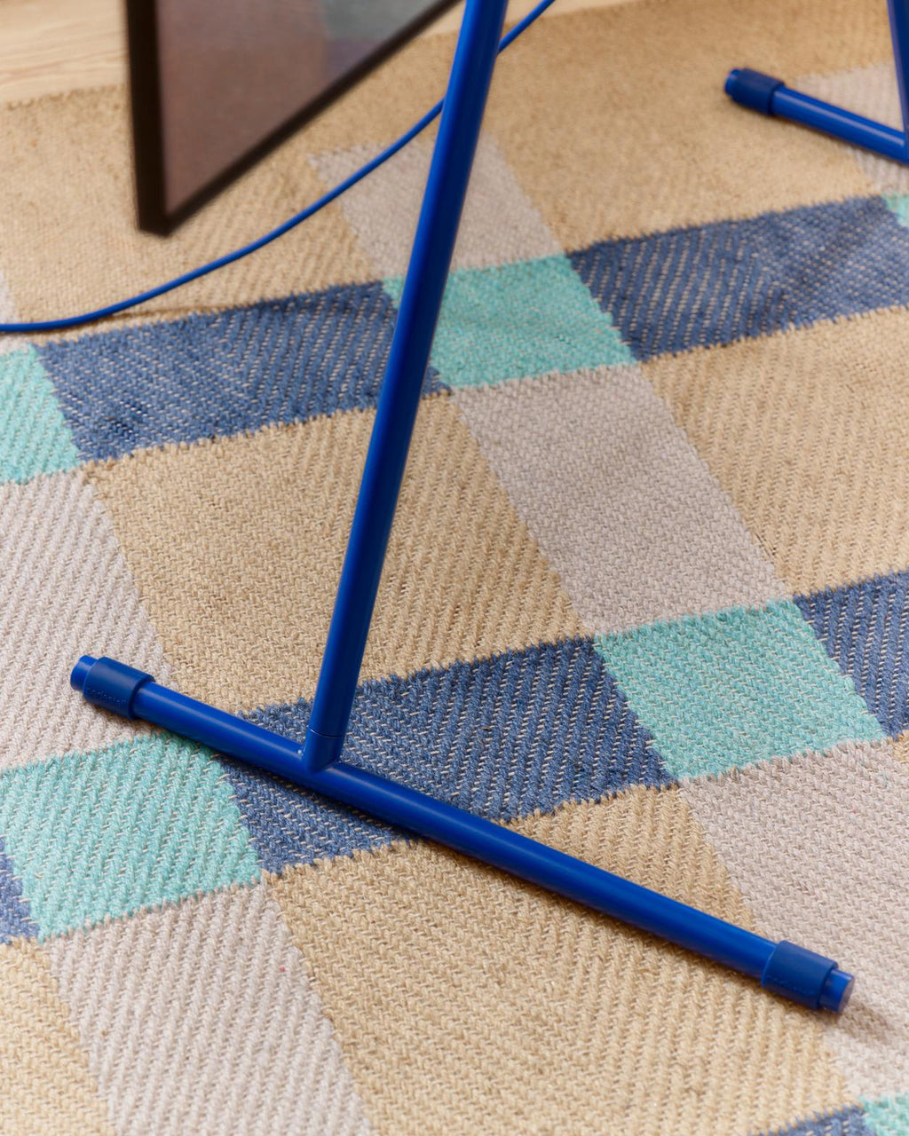 Close-up of the Viva TV stand in Ultra Marine, showing blue tubular feet and cable resting on a checked woven rug