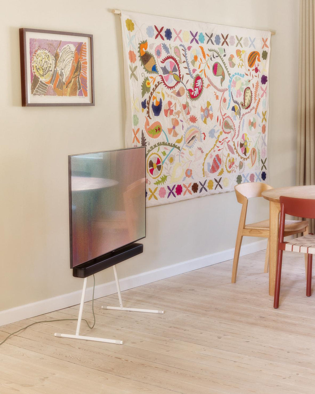 Viva TV Stand in Pearl holding a large TV in a bright dining room with floral wall hanging and oak chairs