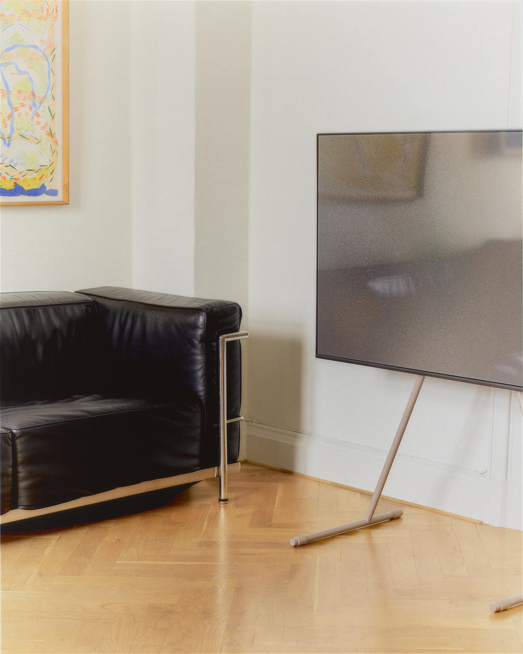 Viva TV stand in Oat holding a large TV in a corner living room with black leather sofa and herringbone wood floor