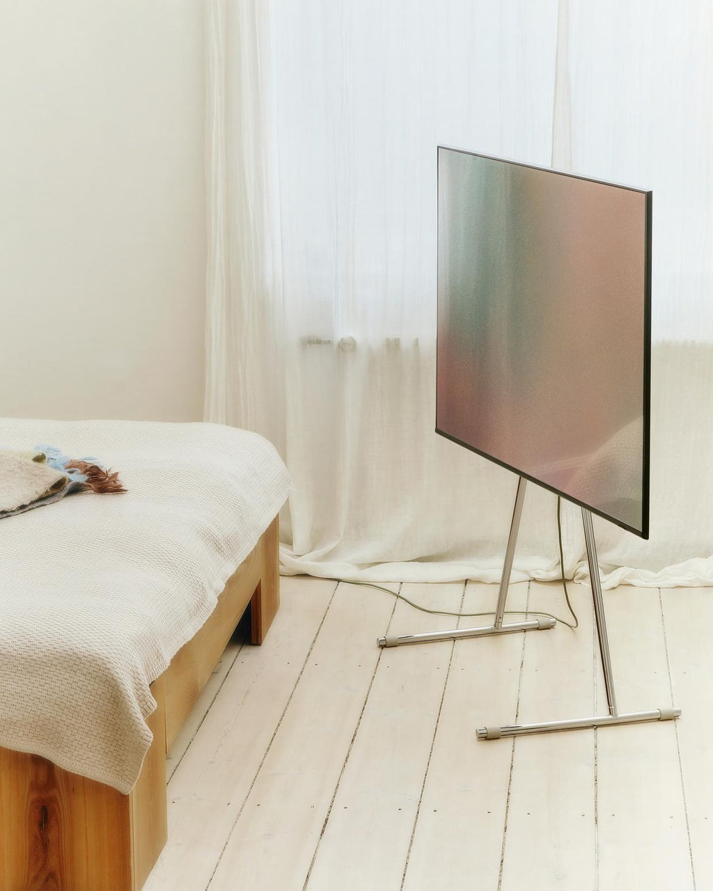 Viva TV stand in chrome holding a large TV in a sunlit bedroom with sheer curtains, pale wood floor and linen bed